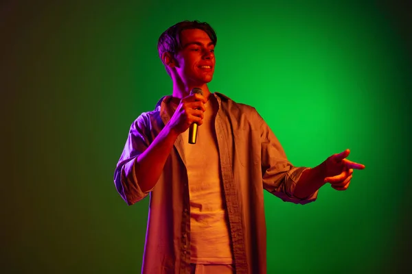 Retrato de joven, cantante pop con micrófono cantando aislado sobre fondo de estudio verde en luz de neón — Foto de Stock
