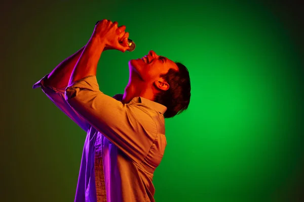 Porträtt av ung man, pop sångare med mikrofon sång isolerad på grön studio bakgrund i neon ljus — Stockfoto