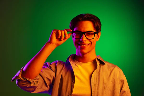 Portret młodego uśmiechniętego mężczyzny umieszczającego okulary na zielonym tle studia w neonie, monochromatyczne. — Zdjęcie stockowe