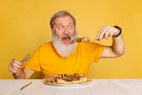 Ενθουσιασμένος γκρίζος γενειοφόρος άνθρωπος τρώει νόστιμη πίτσα με τυρί και ντομάτες που απομονώνονται σε κίτρινο φόντο στούντιο. — Φωτογραφία Αρχείου