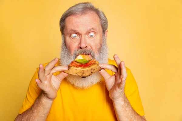 Close-up senior divertido hombre muerde un pedazo de pizza aislado en el fondo del estudio amarillo. Día Mundial de la pizza — Foto de Stock