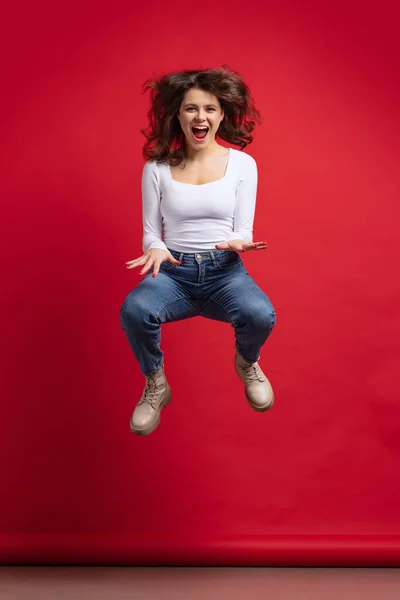 Портрет збудженої молодої красивої дівчини в повсякденному одязі, що стрибає ізольовано на фоні червоного кольору — стокове фото