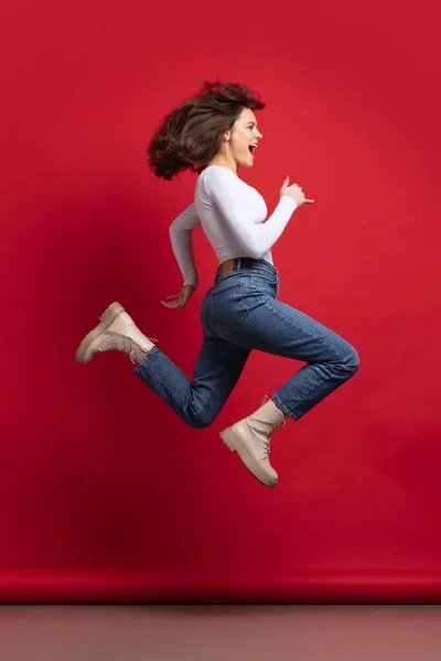 Heyecanlı genç bir kızın dinamik portresi gündelik giysiler içinde kırmızı arka planda izole bir şekilde atlıyor. — Stok fotoğraf