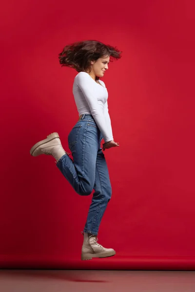 Динамичный портрет возбужденной молодой красивой девушки в повседневной одежде прыжки изолированы на красном фоне — стоковое фото