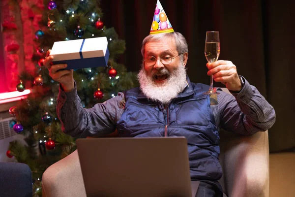 Feliz hombre barbudo viejo sentado en casa y celebra su cumpleaños con amigos y familiares por medio de videollamada. — Foto de Stock