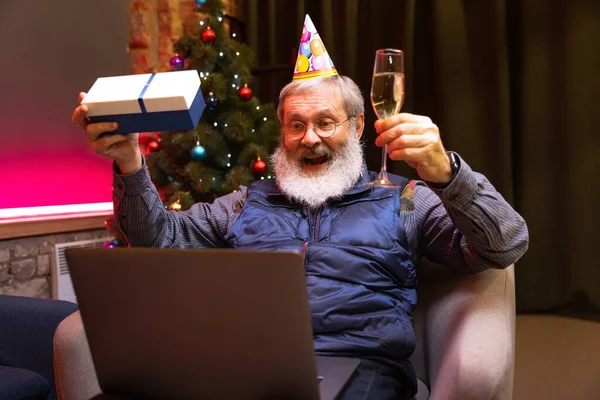 Feliz hombre barbudo viejo sentado en casa y celebra su cumpleaños con amigos y familiares por medio de videollamada. — Foto de Stock