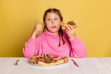 Küçük tatlı bir kızın sarı stüdyo arka planında izole edilmiş lezzetli İtalyan pizzasını tattığı bir stüdyo fotoğrafı..