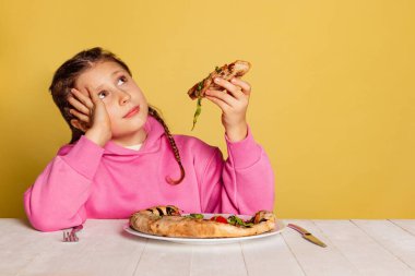 Küçük tatlı bir kızın sarı stüdyo arka planında izole edilmiş lezzetli İtalyan pizzasını tattığı bir stüdyo fotoğrafı..