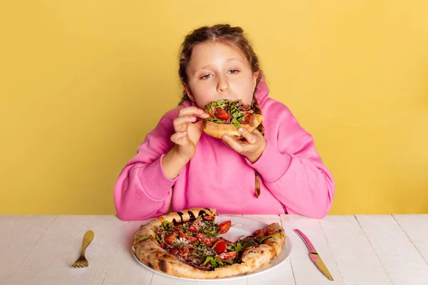 스튜디오에서 작고 귀여운 소녀가 앉아서 노란색 스튜디오 배경에 따로 놓인 맛있는 이탈리아 피자를 맛보는 장면. — 스톡 사진