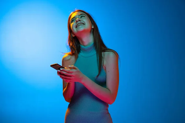 Портрет молодой красивой девушки, студентка, использующая смартфон изолированный на синем фоне в неоновом. Человеческие эмоции, концепция выражения лица — стоковое фото
