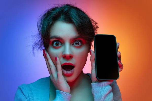 Close-up of shocked, surprised young pretty girl in warm gray knitted sweater with phone isolated on gradient blue orange neon background — 图库照片