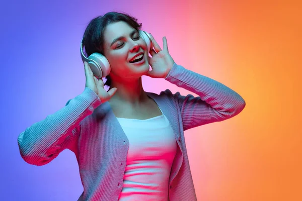 Šťastná mladá hezká dívka v teplé šedé svetr poslech hudby ve sluchátkách izolovaných na gradient modrá oranžová neon pozadí — Stock fotografie