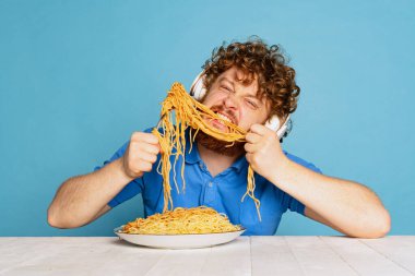 Çılgın genç, kızıl sakallı adam büyük porsiyon erişte tadıyor. Mavi stüdyo arka planında izole edilmiş makarna..