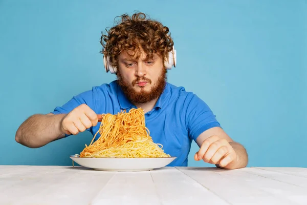 Emotional young hairy red-bearded man tasting large portion of noodles, pasta isolated on blue studio background. — Foto Stock
