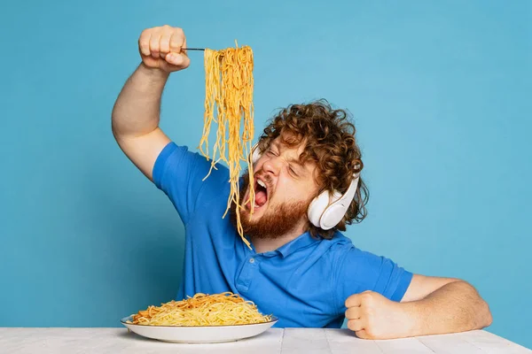 Funny young hairy red-bearded man eating large portion of noodles, pasta isolated on blue studio background.