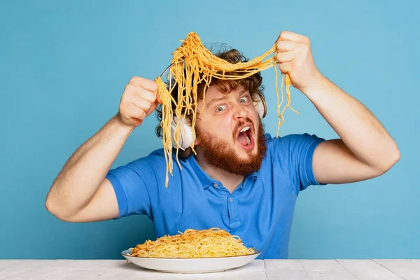 Loco joven peludo hombre de barba roja degustación de gran parte de fideos, pasta aislada sobre fondo de estudio azul. — Foto de Stock