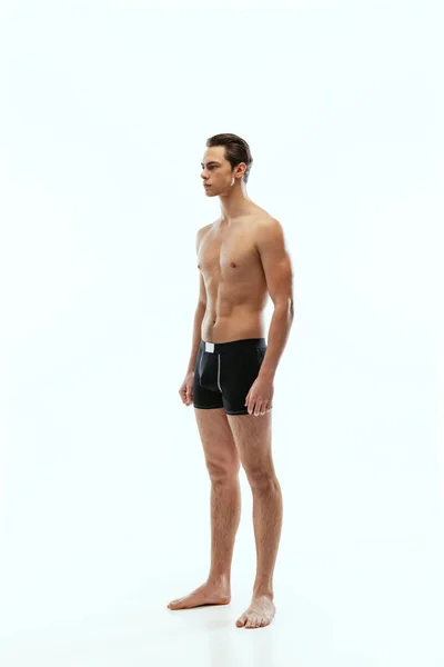 Full-length portrait of young handsome shirtless sportive man wearing black boxer-briefs standing isolated on white studio background. — Stockfoto