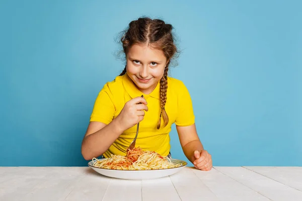 Cute little girl, emotive kid eating delicious Italian pasta isolated on blue studio background. World pasta day — Fotografia de Stock