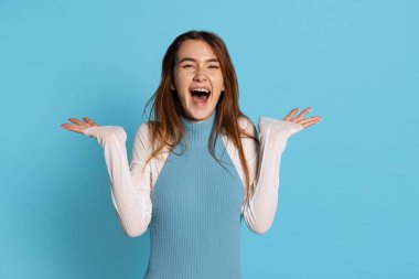 Uzun parlak saçlı güzel kızın yarım boy portresi mavi stüdyo arka planında izole edilmiş zevkle kollarını yana doğru açıyor..