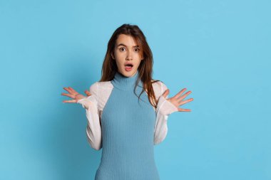 Uzun parlak saçlı güzel bir kızın yarım boy portresi mavi stüdyo arka planında izole edilmiş şok içinde kollarını yana doğru açıyor..