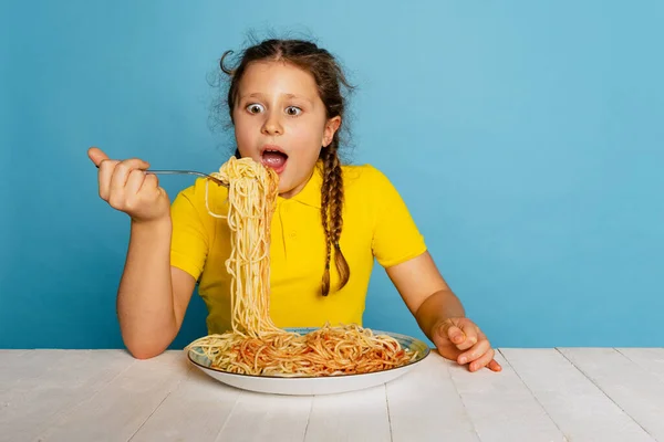 Zszokowana dziewczynka, emocjonujący dzieciak w żółtej koszulce degustacja pysznego włoskiego makaronu odizolowanego na niebieskim tle studia. Światowy dzień makaronu — Zdjęcie stockowe
