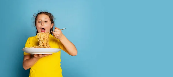 Cute little girl, emotive kid eating delicious Italian pasta isolated on blue studio background. World pasta day — Fotografia de Stock