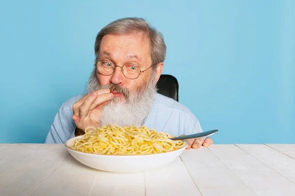 Emotional senior hairy gray-bearded man eating large portion of noodles, pasta isolated on blue studio background. — Stock Photo, Image