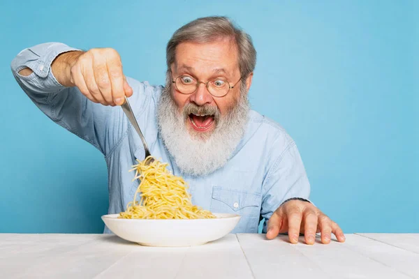 Podekscytowany starszy owłosiony mężczyzna z szarą brodą jedzący dużą porcję makaronu, makaron izolowany na niebieskim tle studia. — Zdjęcie stockowe
