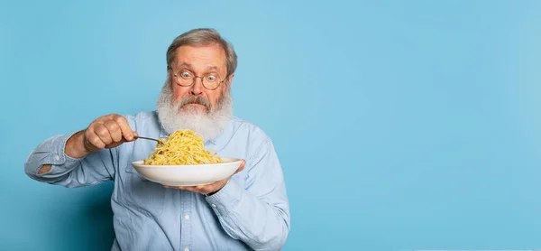 Sorprendido anciano peludo hombre de barba gris comiendo gran parte de fideos, pasta aislada sobre fondo de estudio azul. — Foto de Stock