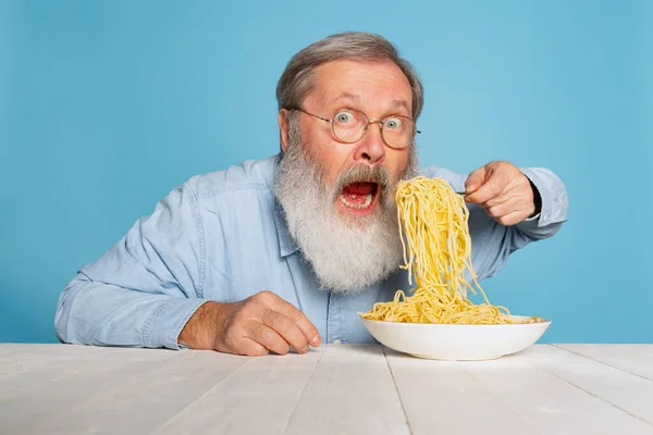 Zszokowany starszy owłosiony mężczyzna z szarą brodą jedzący dużą porcję makaronu, makaron odizolowany na niebieskim tle studia. — Zdjęcie stockowe