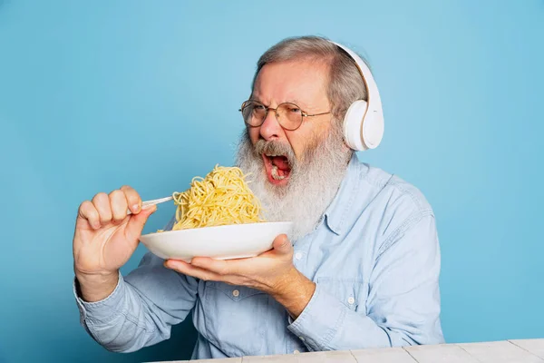 Емоційний старший волохатий сіробородатий чоловік їсть велику порцію локшини, макарони ізольовані на синьому студійному фоні . — стокове фото