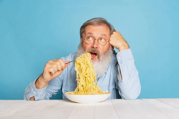Duygusal son sınıf kıllı, gri sakallı adam büyük porsiyon erişte yiyor. Mavi stüdyo arka planında izole edilmiş makarna.. — Stok fotoğraf