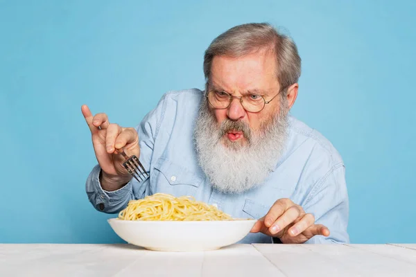 Fastidioso sénior peludo hombre de barba gris degustación de gran porción de fideos, pasta aislada sobre fondo de estudio azul. — Foto de Stock