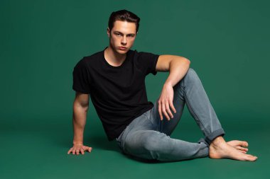 Stüdyoda, siyah tişört ve ekose gömlek giyen yakışıklı bir adamın yeşil stüdyo arka planında izole bir şekilde duran kot pantolonu..