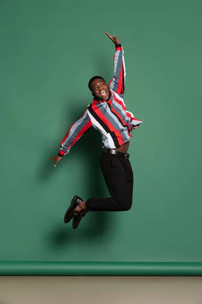 Studyjne ujęcie afrykańskiego mężczyzny w jasnej koszuli skaczącego na ciemnozielonym tle. Wow koncepcja emocji — Zdjęcie stockowe