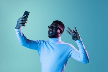 Beyaz yaka polo ve kot giyen, mavi arka planda izole edilmiş telefon kullanan, neon ışıklı genç bir öğrencinin stüdyo fotoğrafı..