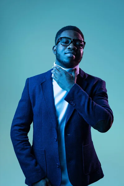 Half-length portrait of young stylish african man, student wearing white neck-polo and jacket isolated on blue background in neon light. — Fotografia de Stock