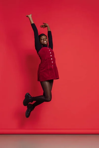 Ganztägiges Porträt eines glücklichen afrikanischen Mädchens in lässiger Kleidung, das isoliert auf rotem Studiohintergrund springt. Konzept der Emotionen, Urlaub — Stockfoto