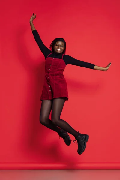 Full-length portrait of happy African girl in casual clothes jumping isolated on red studio background. Concept of emotions, holidays — Stockfoto