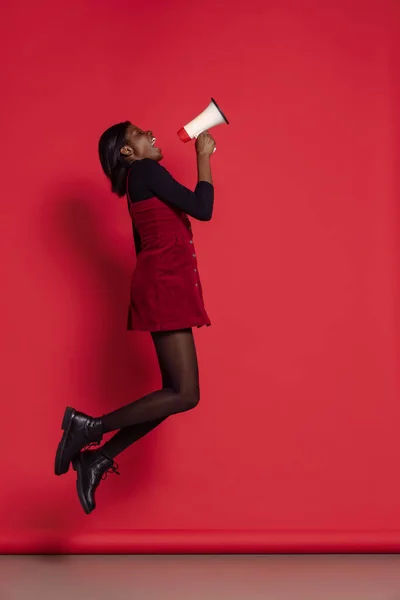 Retrato de larga duración de la chica africana feliz en ropa casual saltando aislado en el fondo del estudio rojo. Concepto de emociones, vacaciones — Foto de Stock
