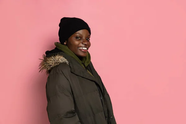 Sonriente chica africana joven con ropa de abrigo de invierno y sombrero de pie solo aislado en el fondo del estudio rosa con copyspace para el anuncio. — Foto de Stock