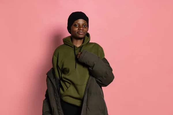 Smiling young african girl wearing winter warm clothes and hat standing alone isolated on pink studio background with copyspace for ad. — Stockfoto