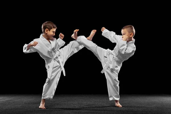 Dos niños pequeños, chicos, atletas de taekwondo entrenando juntos aislados sobre un fondo oscuro. Concepto de deporte, educación, habilidades — Foto de Stock