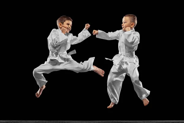 Δύο μικρά παιδιά, αγόρια, αθλητές taekwondo προπονούνται μαζί απομονωμένοι σε σκοτεινό φόντο. Έννοια του αθλητισμού, της εκπαίδευσης, των δεξιοτήτων — Φωτογραφία Αρχείου