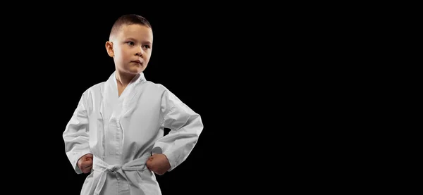 Um miúdo, rapaz, atleta de taekwondo a usar dobok branco isolado sobre fundo escuro. Conceito de esporte, educação, habilidades — Fotografia de Stock