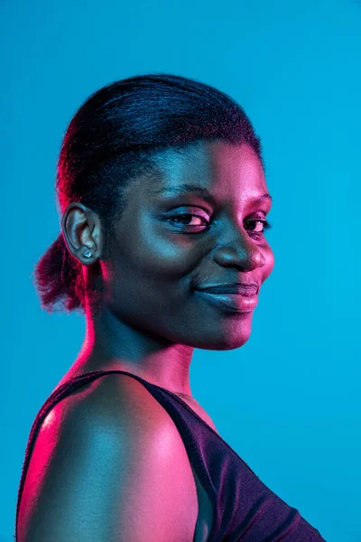 Close-up of young beautiful dark skinned woman posing isolated on blue studio background in neon light. — Stockfoto