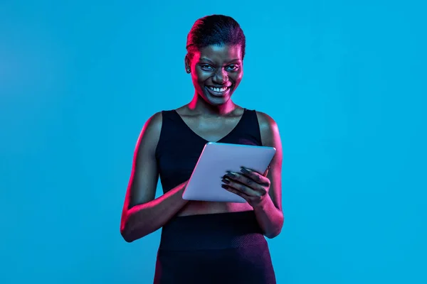 Portrét mladé krásné tmavé kůže žena pomocí tabletu izolované na modrém pozadí studia v neonovém světle. — Stock fotografie