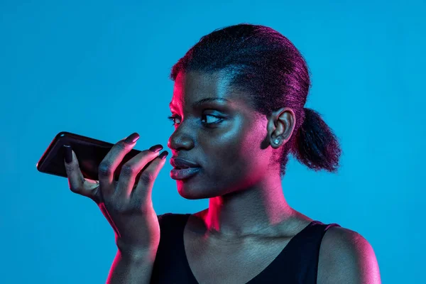 Retrato de una joven hermosa mujer de piel oscura utilizando el teléfono aislado en el fondo del estudio azul en luz de neón. — Foto de Stock