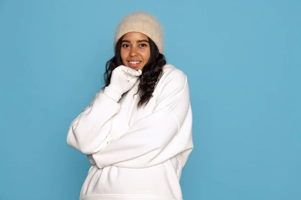 Smiling young beautiful dark skinned girl in warm white hoodie posing isolated on blue background. — Fotografia de Stock