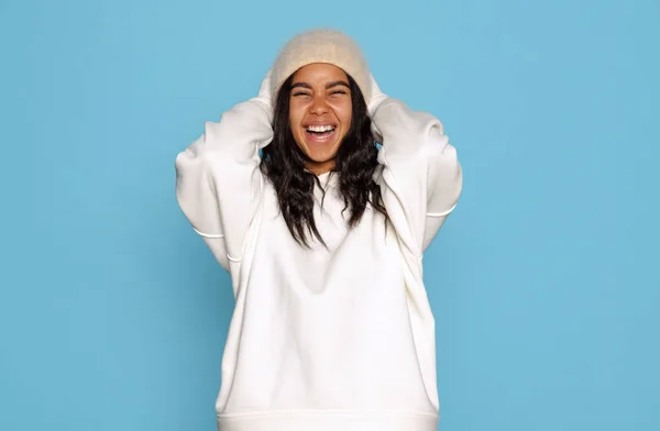 Happy, excited young beautiful dark skinned girl in warm white hoodie and hat isolated on blue background. — Stock Photo, Image